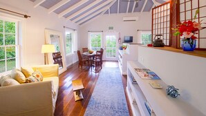 Une pièce spacieuse avec un plafond haut, des poutres apparentes, un coin repas avec des chaises et une table, un canapé et un téléviseur posé sur une étagère.