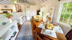 Un coin repas avec une table en bois dressée pour quatre personnes, une fenêtre donnant sur un paysage verdoyant et un ventilateur de plafond.