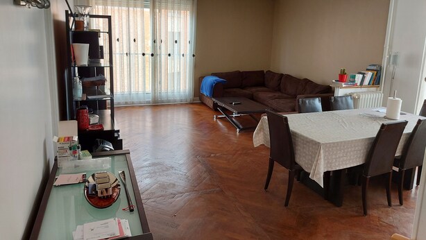 Een woonkamer met een bruine leren bank, een eettafel met stoelen, een glazen salontafel en een boekenkast.
