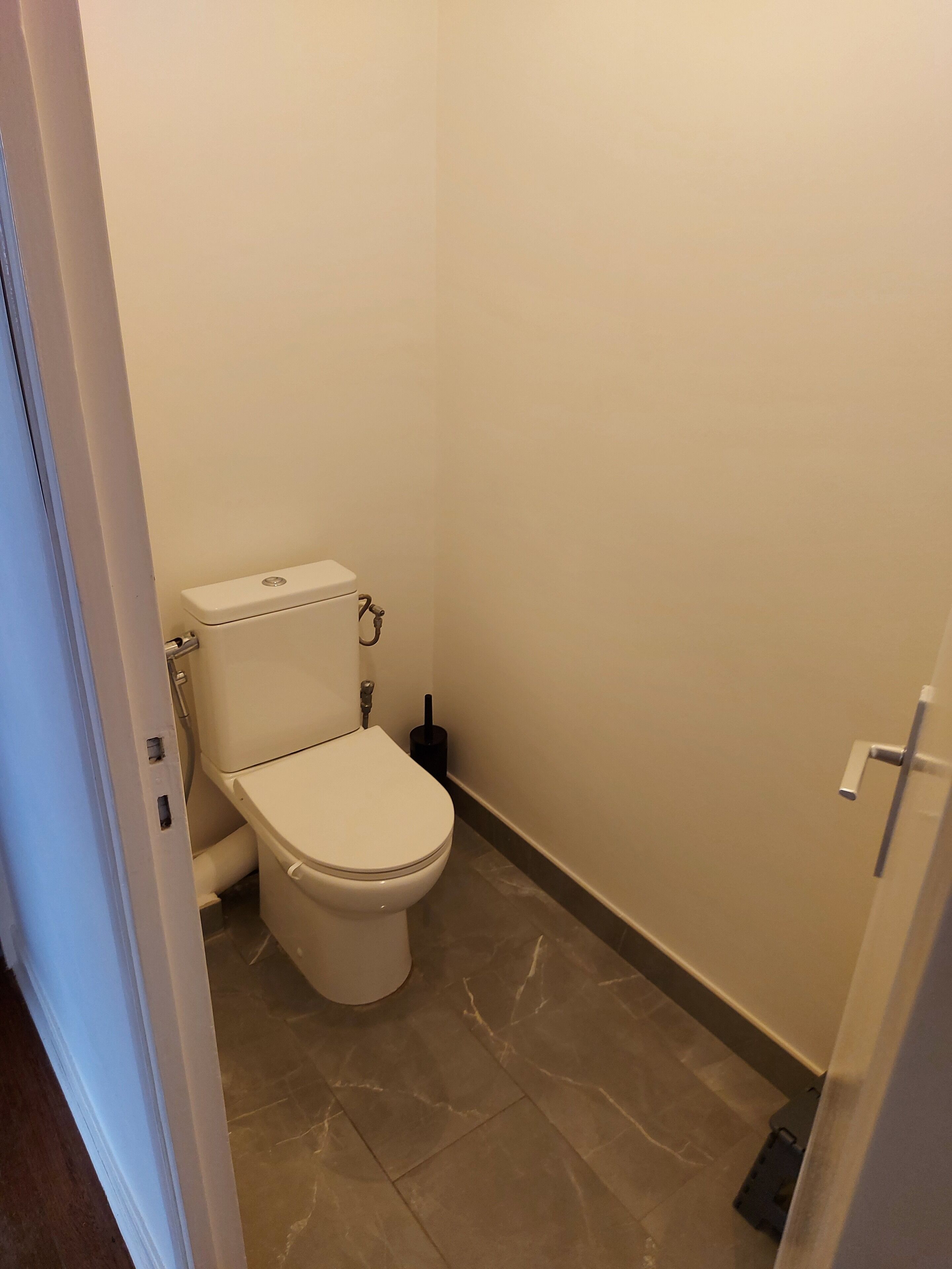 A small, clean bathroom with a toilet, a wall-mounted flush handle, and a floor drain.