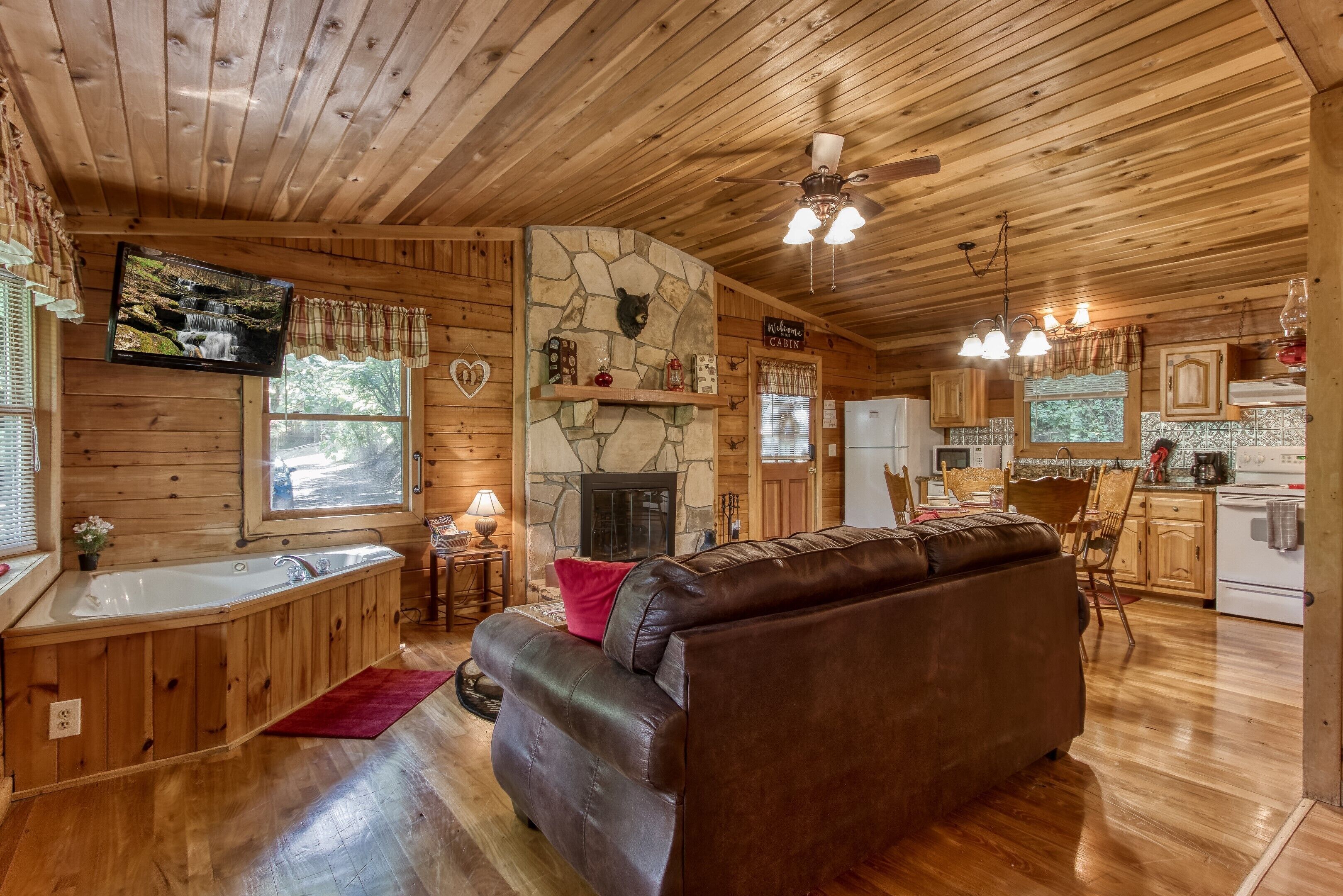 A cabin interior with a stone fireplace, wooden walls, a leather sofa, a kitchen area, and a large window.
