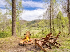 A campfire with two Adirondack chairs in a forested area with a view of rolling hills.