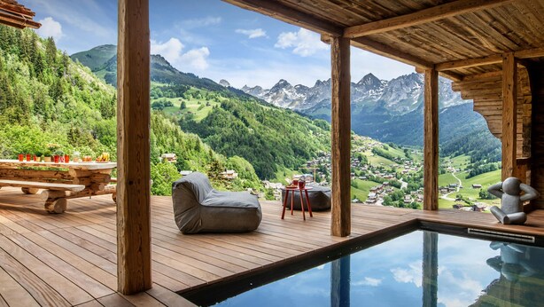 A wooden deck with a pool, outdoor seating, and a scenic mountain view. - Chalet Happyview - luxury holiday for 15 with pool & sauna - OVO Network (Le Grand-Bornand)