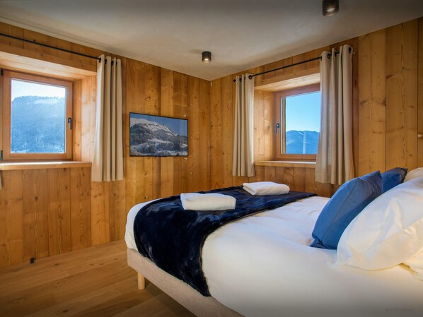 Un dormitorio con paneles de madera, dos ventanas, una cama con ropa de cama blanca y azul, y un televisor en la pared.