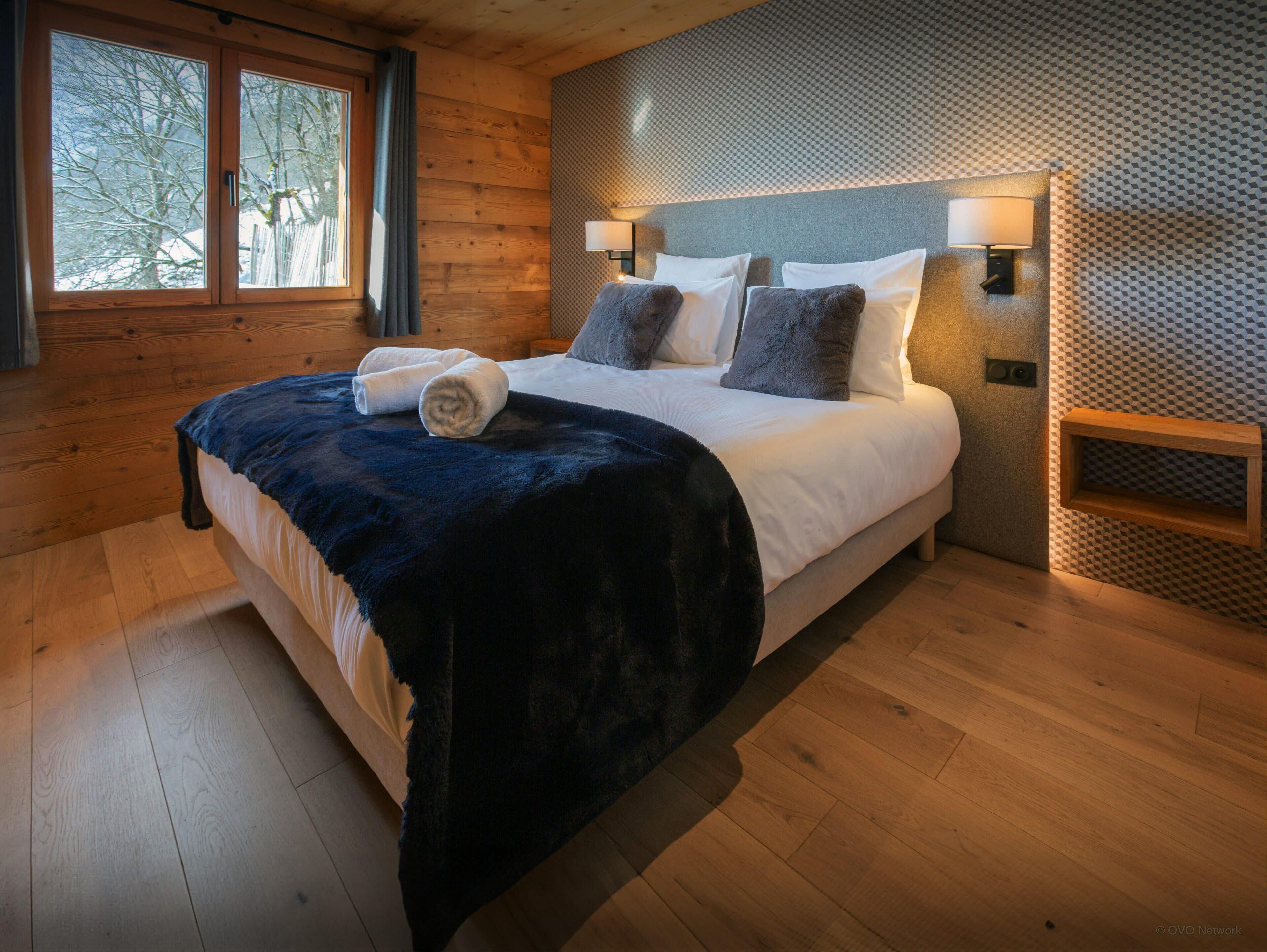 A bedroom with a large bed, wooden walls, and a window with a view of trees.