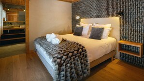 A bedroom with a wooden ceiling, a textured wall, a bed with white bedding, a fur throw, and a bedside table with a lamp. - Chalet Happyview - luxury holiday for 15 with pool & sauna - OVO Network (Le Grand-Bornand)