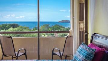 A balcony with a view of the ocean and a small island. There are two chairs and a bed with a patterned bedspread.