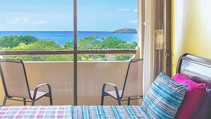 A balcony with two chairs and a table, overlooking a coastal view with greenery and the sea.