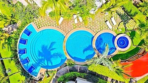 A large outdoor swimming pool with multiple levels, surrounded by lush greenery and palm trees.