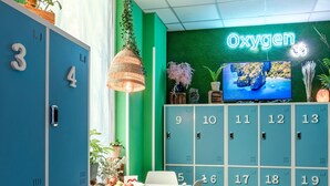 A room with blue lockers numbered 1-19, a white table with chairs, a hanging lamp, and a neon sign reading "Oxygen". - Oxygen Hostel (Madrid)