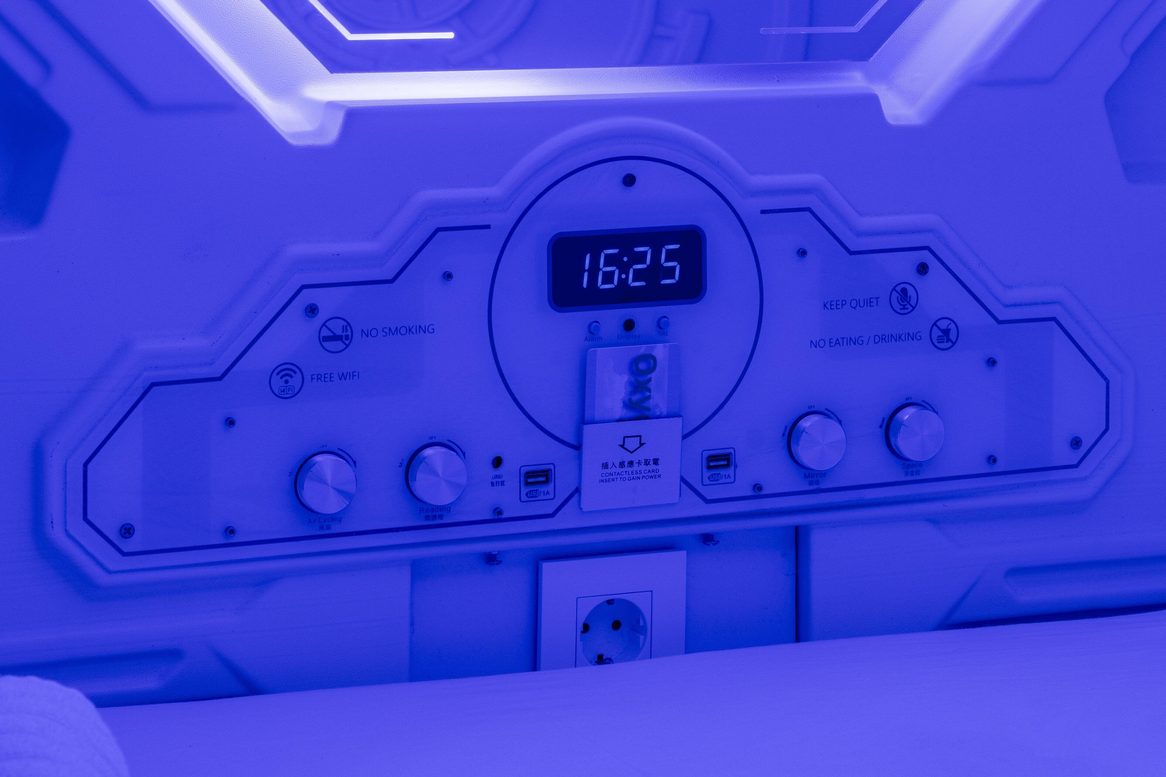 A control panel with a digital clock displaying 16:25, various buttons, and labels indicating no smoking, free wifi, and no eating/drinking.