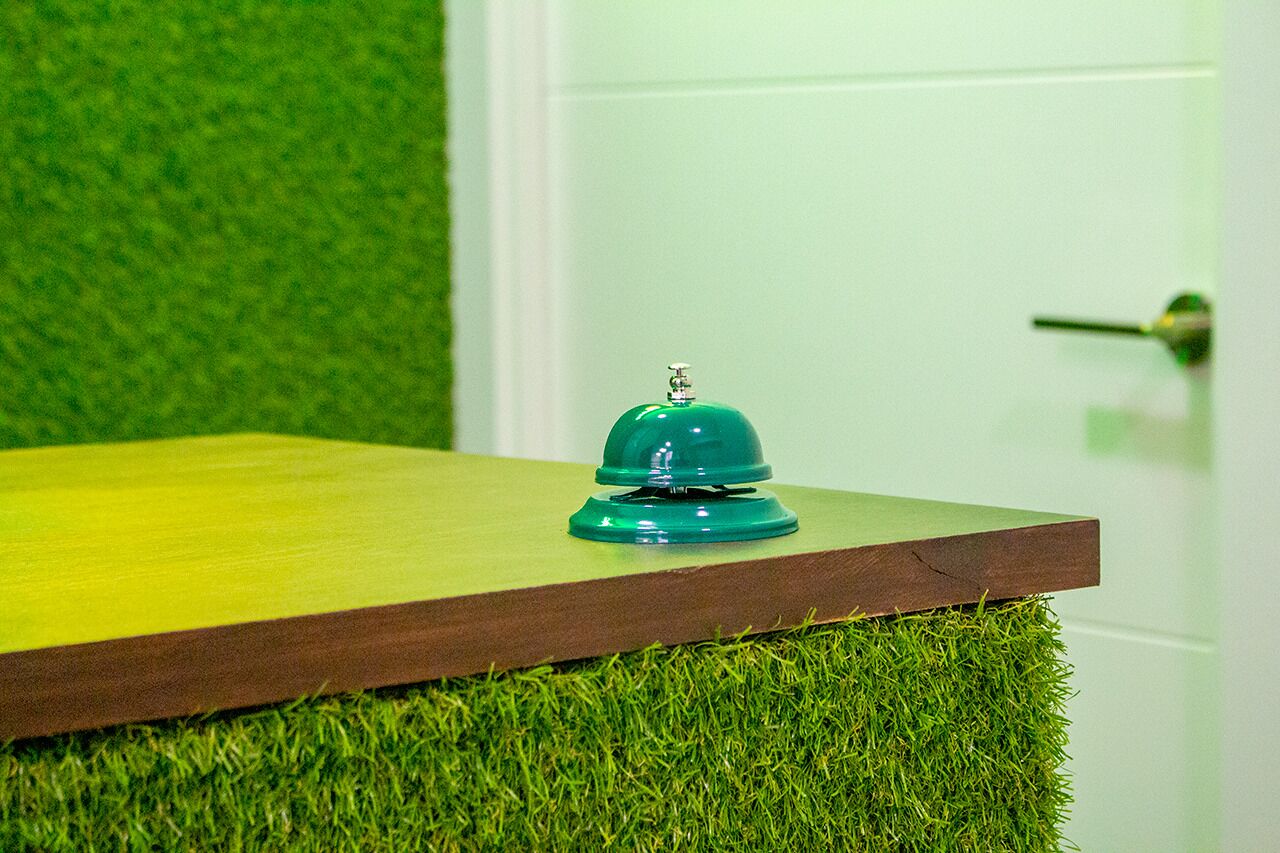 Uma mesa de madeira com um sino verde em cima, com grama sintética e uma parede verde ao fundo.