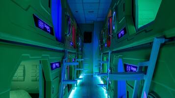 Un couloir futuriste, illuminé de néons, avec des lits superposés et des fenêtres rondes.