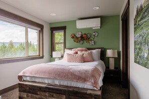 A bedroom with a large bed, two bedside tables with lamps, a wall-mounted air conditioner, and a large window with a view of trees.