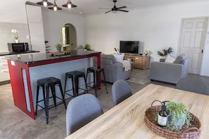 A modern living room with a kitchen area, grey sofas, a wooden dining table, and a TV.