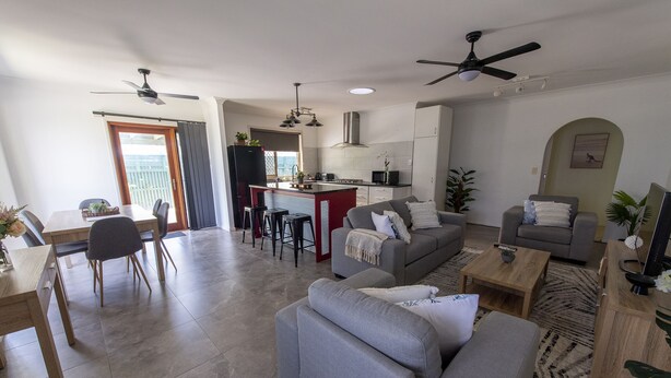 A modern living room with a grey sofa, a dining area with a table and chairs, a kitchen with a central island, and a ceiling fan. - Pet Friendly, 2 Bedroom, Sleeps 4, 250m to Beach, Close to Cafes, Sunshine Coast (Marcoola)