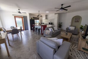 A modern living room with a grey sofa, a dining area with a table and chairs, a kitchen with a central island, and a ceiling fan.