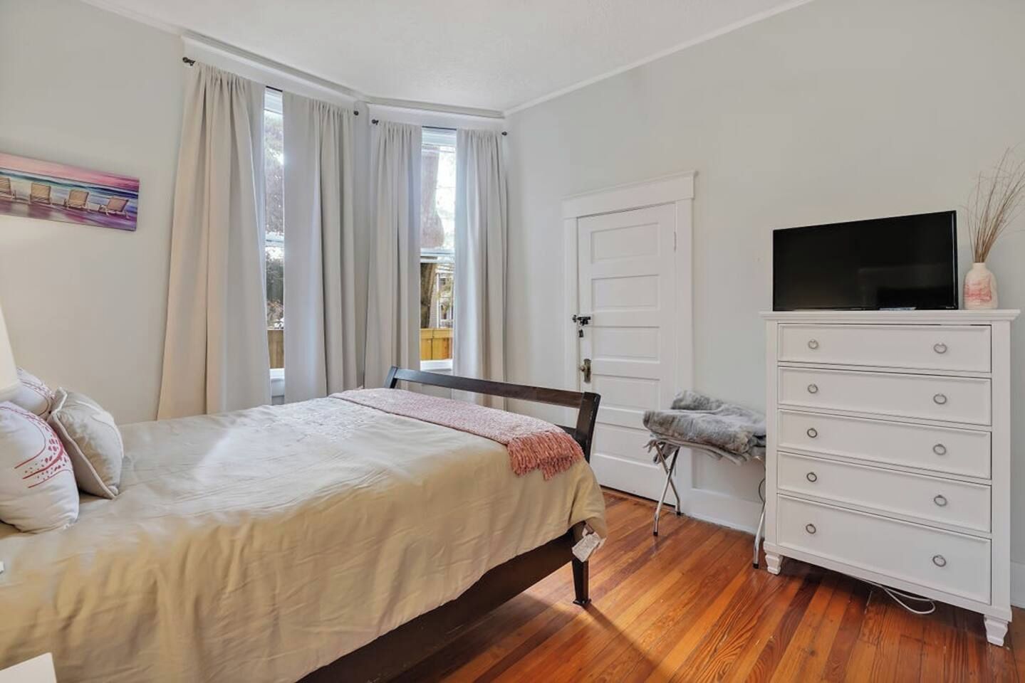 Une chambre à coucher avec un lit, une commode, un téléviseur et une fenêtre avec des rideaux.