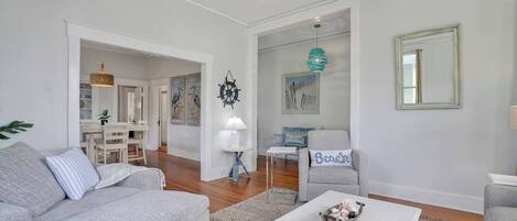 Una sala de estar moderna con un sofá gris, una mesa de centro blanca y un sillón gris. Se observa un comedor al fondo con una mesa y sillas.