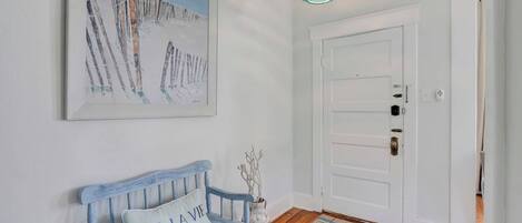 A hallway with a white door, a blue bench with a cushion, a painting, and a hanging light fixture.