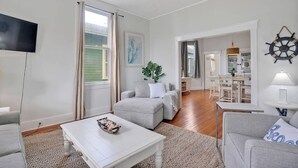 Ein geräumiges Wohnzimmer mit Flachbildfernseher, grauen Sofas, einem weißen Couchtisch und einem großen Fenster mit Vorhängen.