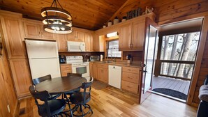 A wooden cabin kitchen with a dining table, chairs, a refrigerator, microwave, stove, and a window with a view of trees.
