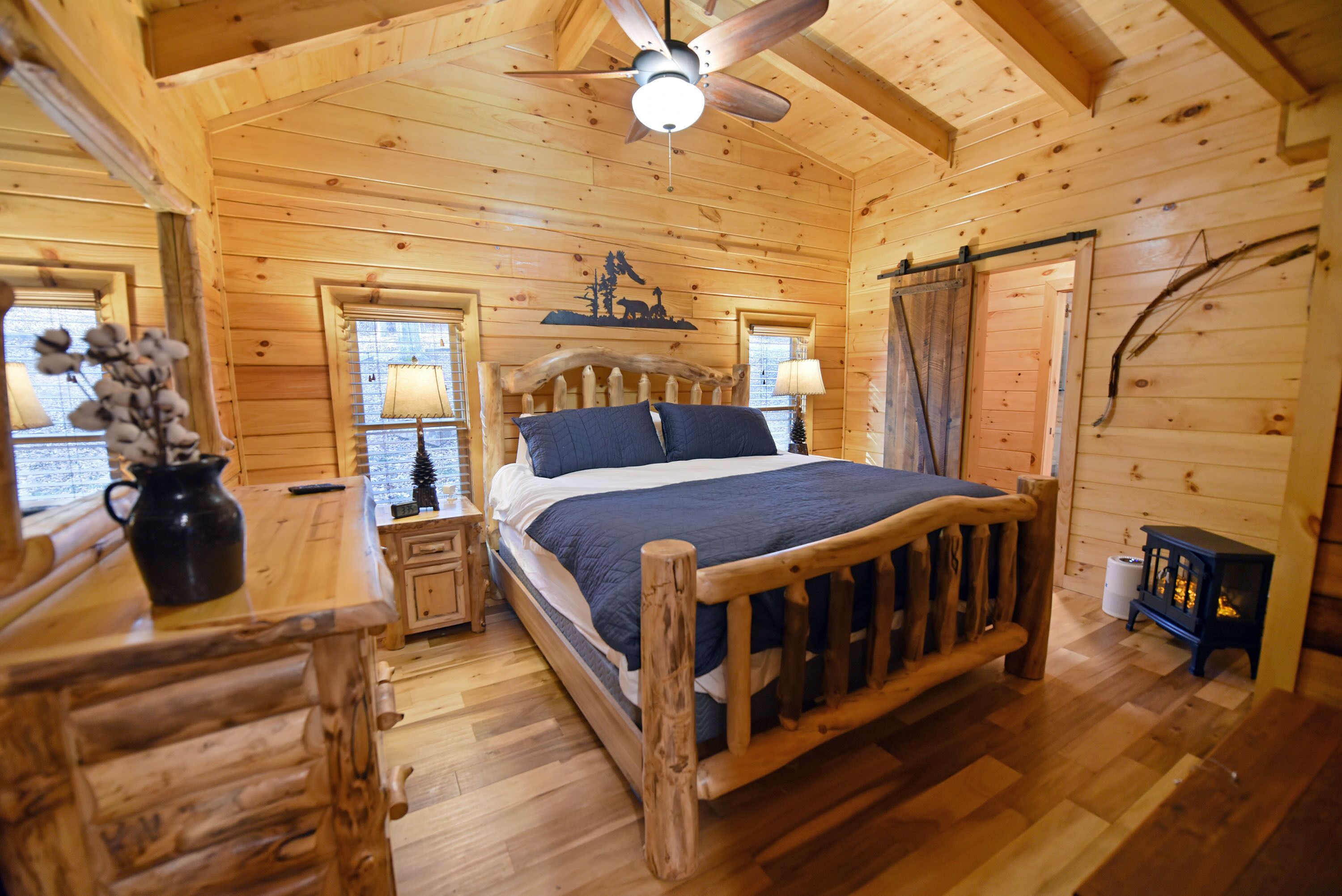 Une chambre à coucher dans une cabane en bois, avec un lit, une table de chevet, une lampe, une cheminée et un ventilateur de plafond.