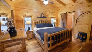 A wooden cabin bedroom with a bed, a nightstand, a lamp, a fireplace, and a ceiling fan.