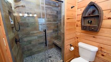 A wooden cabin bathroom with a glass-enclosed shower, a wooden shelf with a camera, and a white toilet.