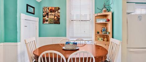 A dining area with a wooden table and white chairs, a white door, and a window with blinds.