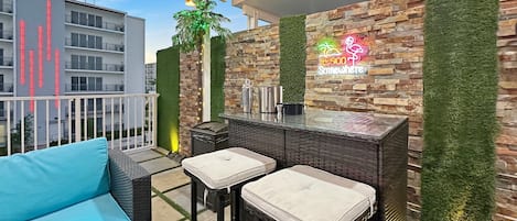 A balcony with wicker furniture, a bar area with a neon sign, and a view of a building with vertical light strips.