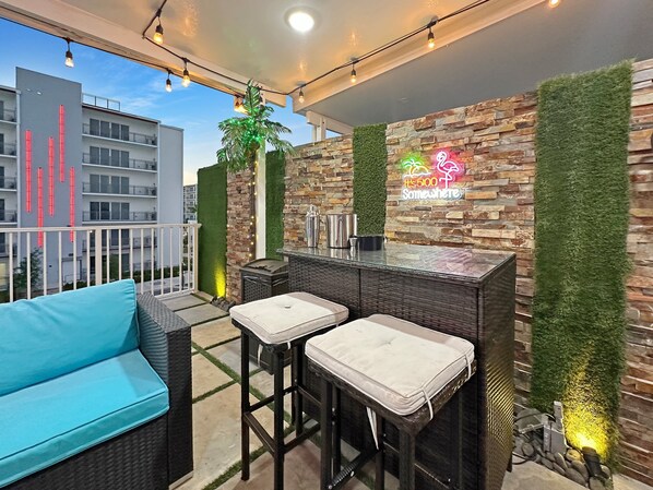 A balcony with wicker furniture, a bar area with a neon sign, and a view of a building with vertical light strips.