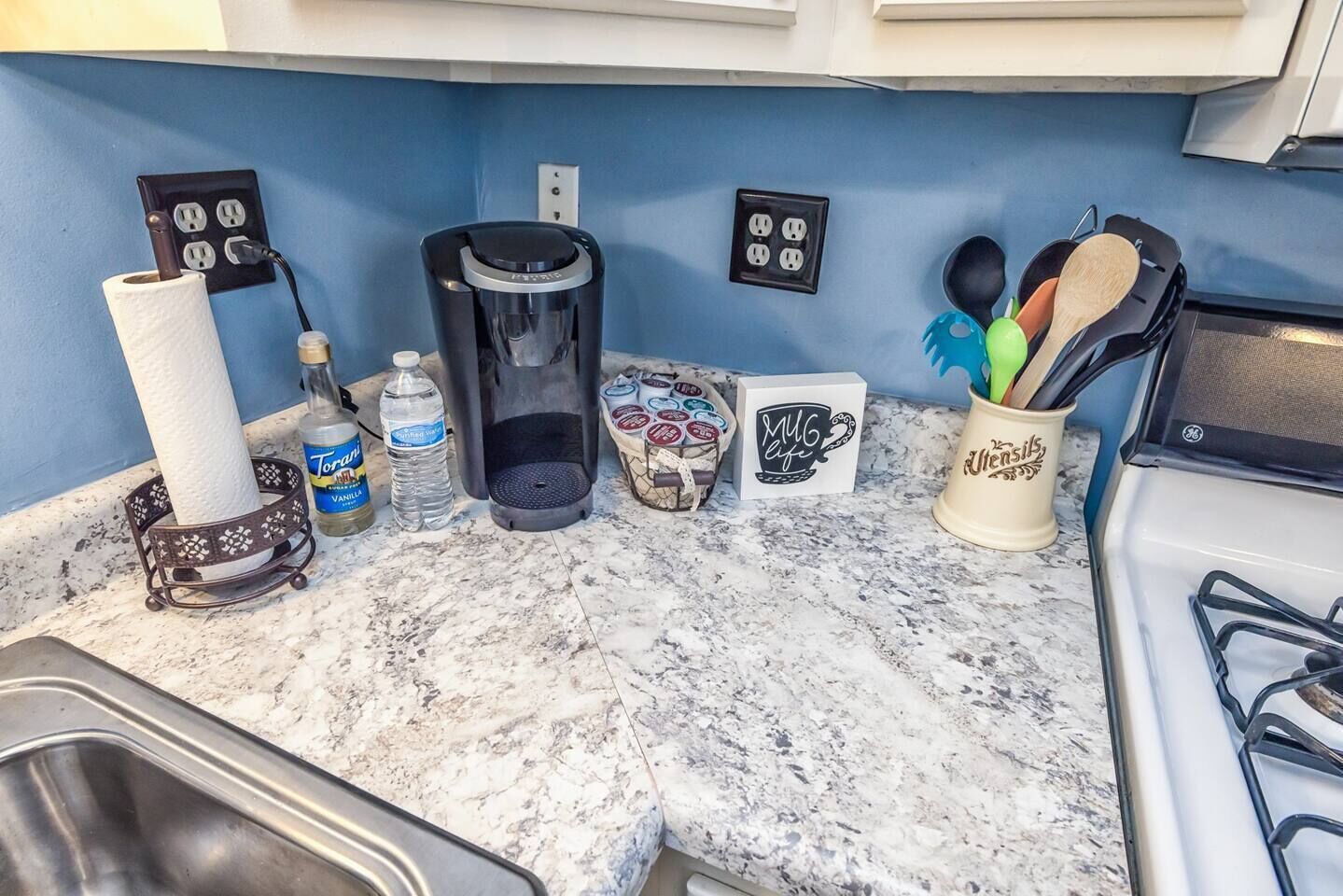 A kitchen counter with a coffee maker, paper towel holder, water bottle, and a mug with utensils.
