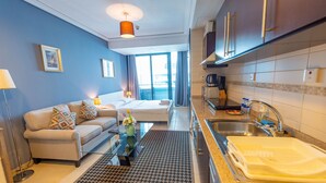 A modern hotel room with a sofa, a coffee table, a kitchen area with a sink and stove, and a view of the city through large windows.