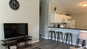 Ein modernes Wohnzimmer mit Flachbildfernseher, Couchtisch und Sofa. Es gibt eine Küchenzeile mit Barhocker und Kühlschrank.