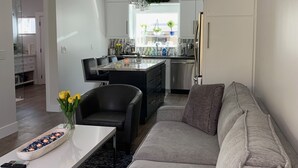 A modern living room with a grey sofa, a dining table with yellow flowers, and a kitchen in the background.