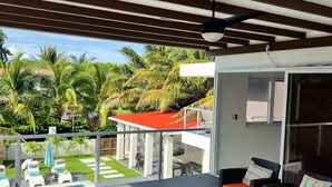 A covered outdoor seating area with wicker furniture and orange cushions, a pool, and a view of palm trees and a building in the background. - Lazo Beach House, Costa del Sol (San Luis, La herradura)