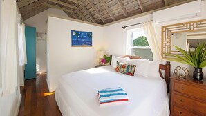 A bedroom with a wooden ceiling, a bed with white bedding, a blue and white striped pillow, a wooden dresser, and a window with a view of greenery.