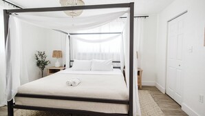 A four-poster bed with white bedding, a wooden floor, a small bedside table with a lamp, and a plant.