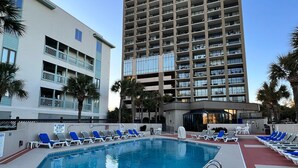 A hotel with a swimming pool, lounge chairs, and a tall building in the background. - Oceanfront Condo w/ Balcony - King Bed - Myrtle Beach - Pool (Myrtle Beach)