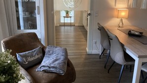 Uma sala de estar moderna com um sofá de couro marrom, um cobertor cinza e uma pequena planta em vaso. Uma mesa de jantar de madeira com cadeiras é visível ao fundo.