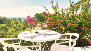 A round white table with two chairs, a bottle of wine, and two glasses on a deck with a view of a garden and ocean. - 1BR Hillside Cottage on Peaceful Private Estate (Governor's Harbour)