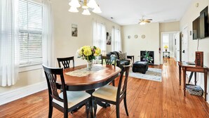 Uma sala de jantar com uma mesa de madeira, cadeiras e um vaso de flores. Há também uma sala de estar com um sofá e uma televisão.