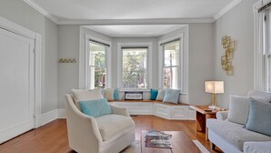 A living room with a beige sofa, a wooden coffee table, and a white armchair. There are windows with white frames and a view of trees outside. - Savannah Suite on Front (Wilmington)