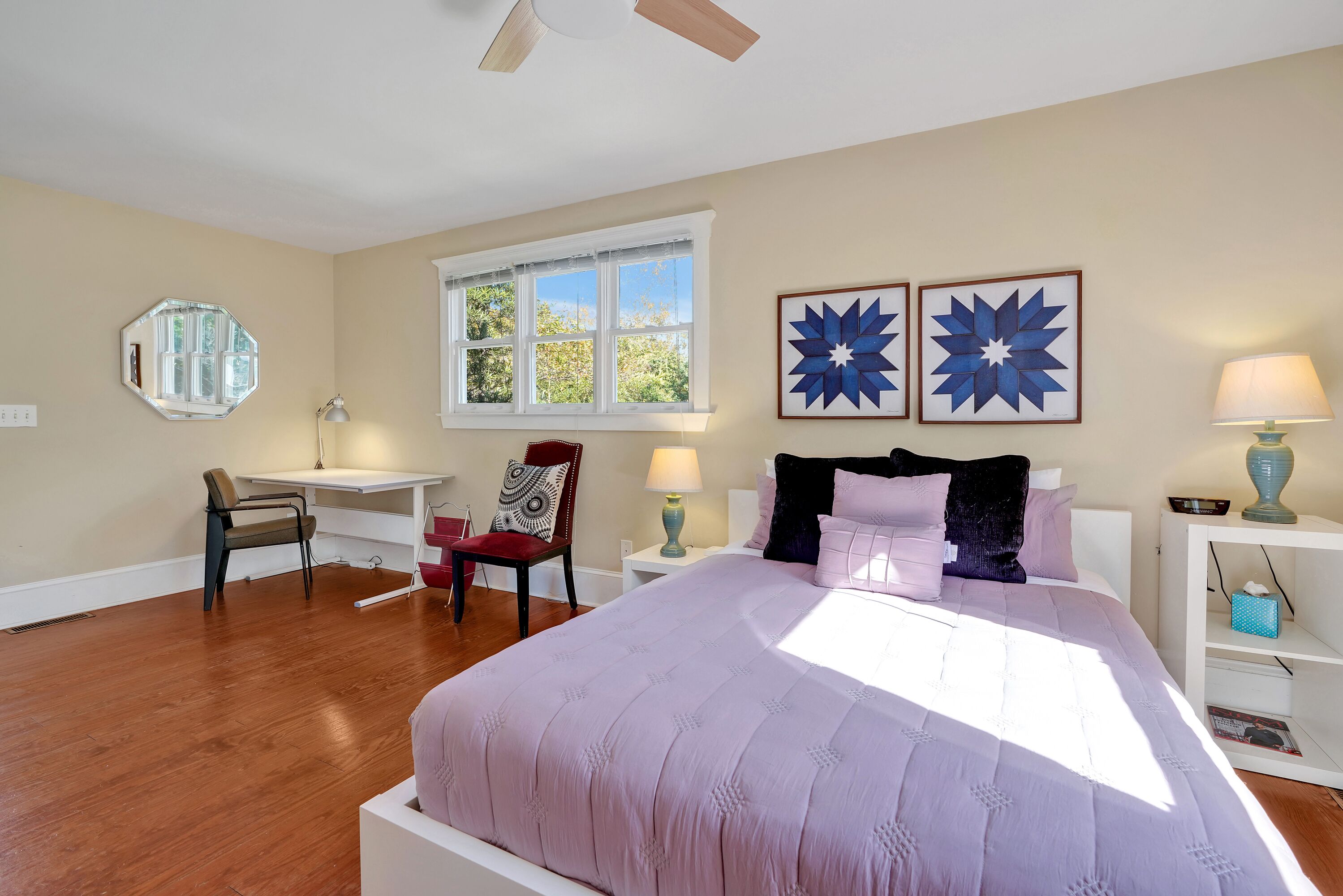 A bedroom with a large bed, a desk, a chair, a window, and artwork on the wall.