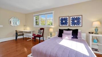 A bedroom with a large bed, a desk, a chair, a window, and artwork on the wall.