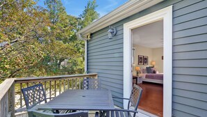 A wooden deck with a table and chairs, a door leading to a room with a bed and a nightstand, and a view of trees. - Condo Riviere Stay Downtown! Walk everywhere! (Wilmington)