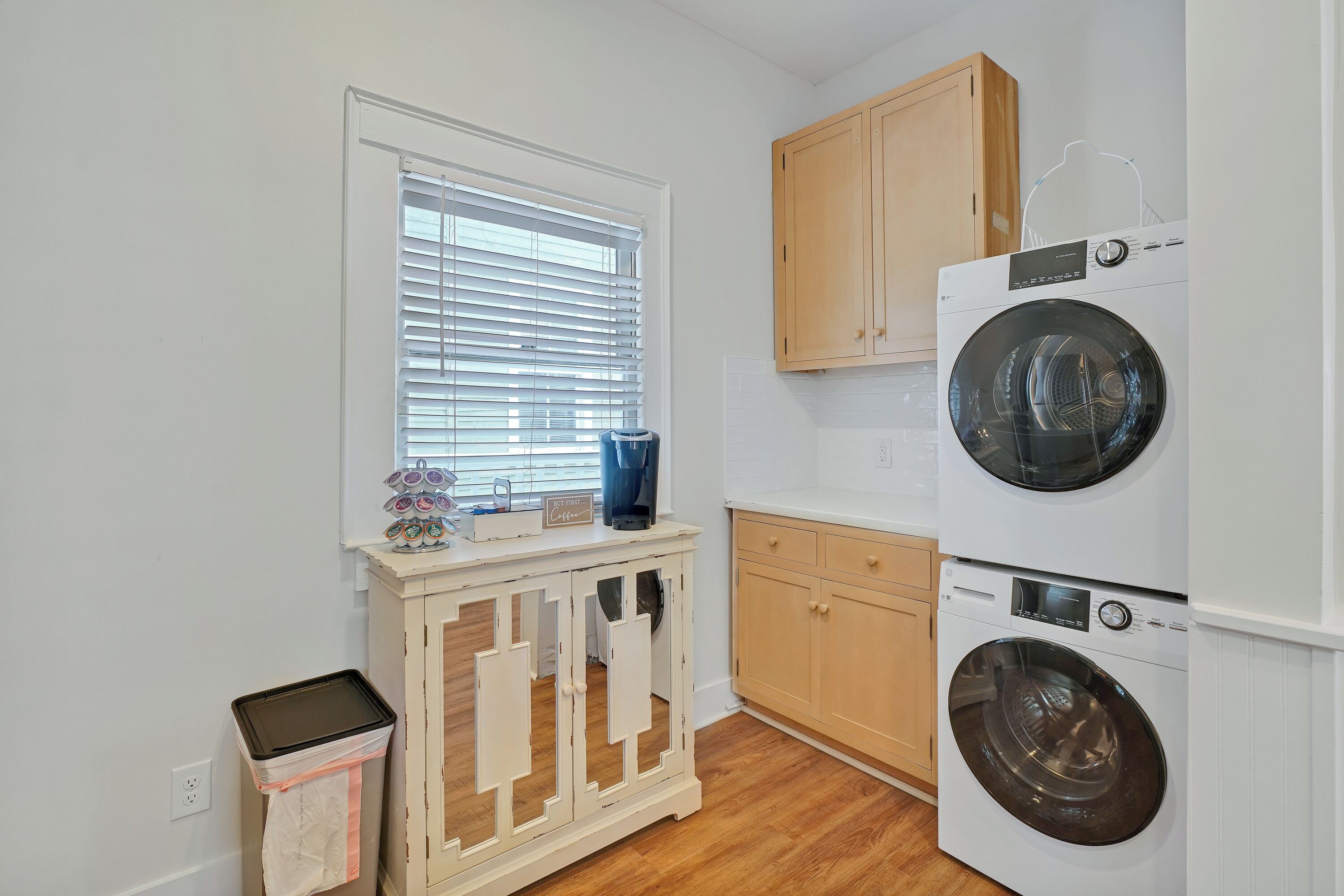 A laundry room with two stacked front-loading washing machines, wooden cabinets, a window with blinds, and a small trash bin.