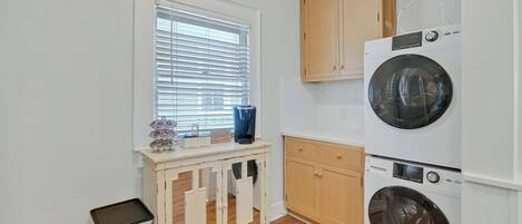 A laundry room with two stacked front-loading washing machines, wooden cabinets, a window with blinds, and a small trash bin.