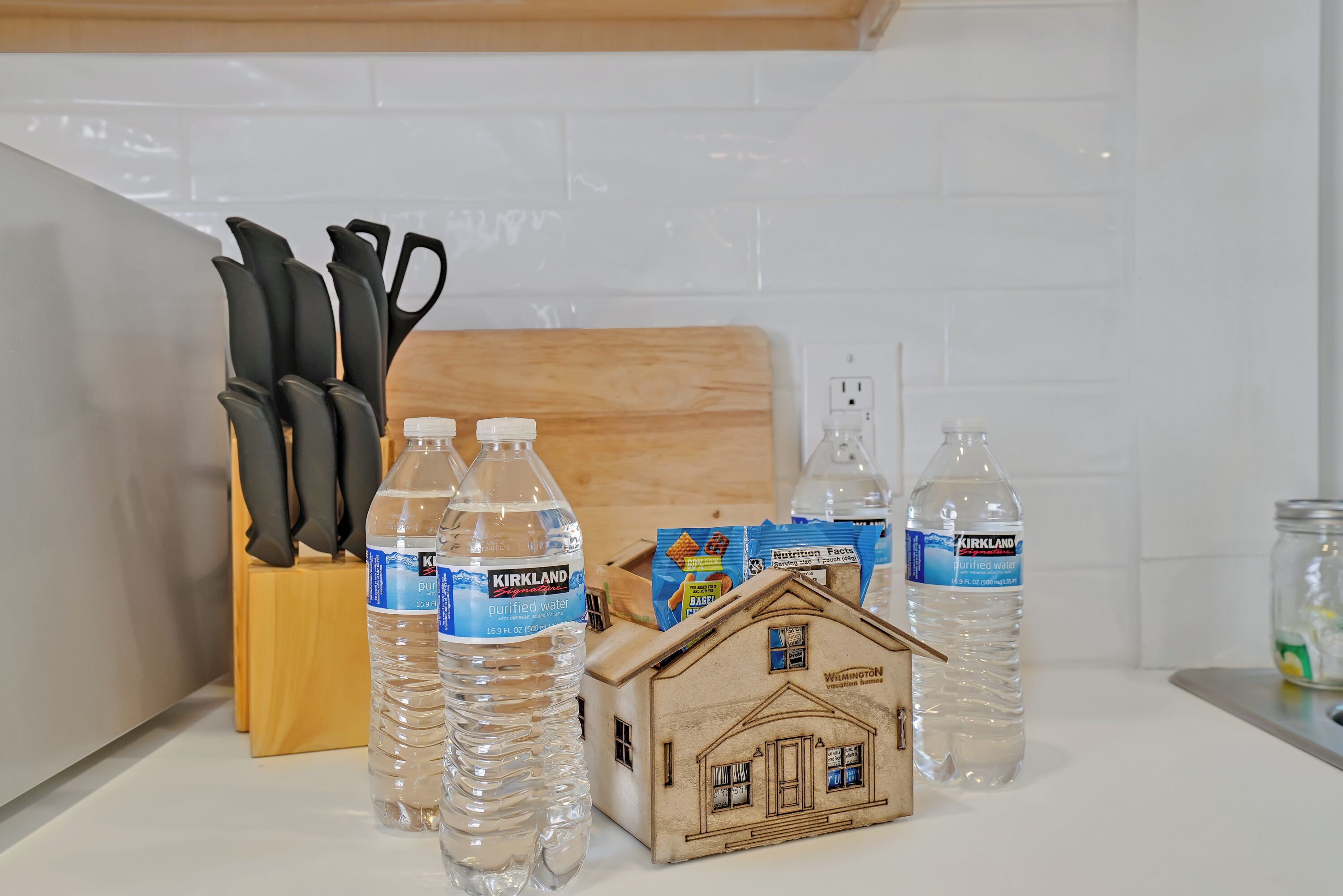 Un comptoir de cuisine avec des bouteilles d’eau de la marque Kirkland, une maquette de maison en bois et un bloc à couteaux.